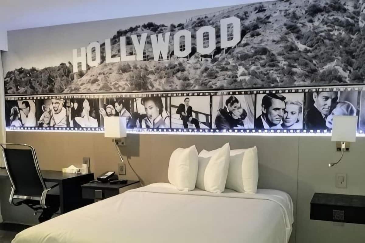A hotel room with a large Hollywood-themed wall, a bed with white bedding, a desk with a chair, and a nightstand with a lamp.