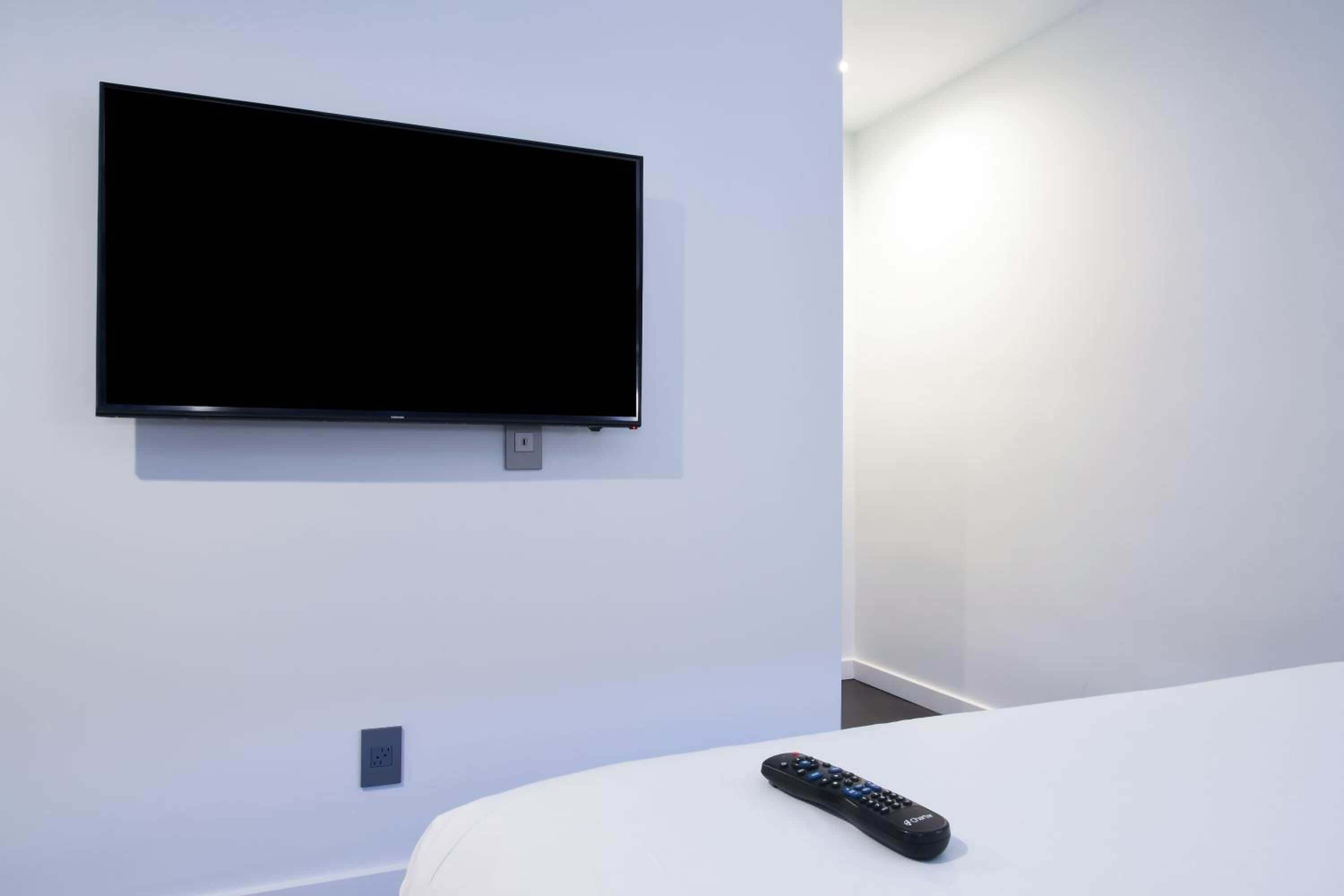 A modern hotel room with a flat-screen TV mounted on the wall, a remote control on the bed, and a minimalist design.