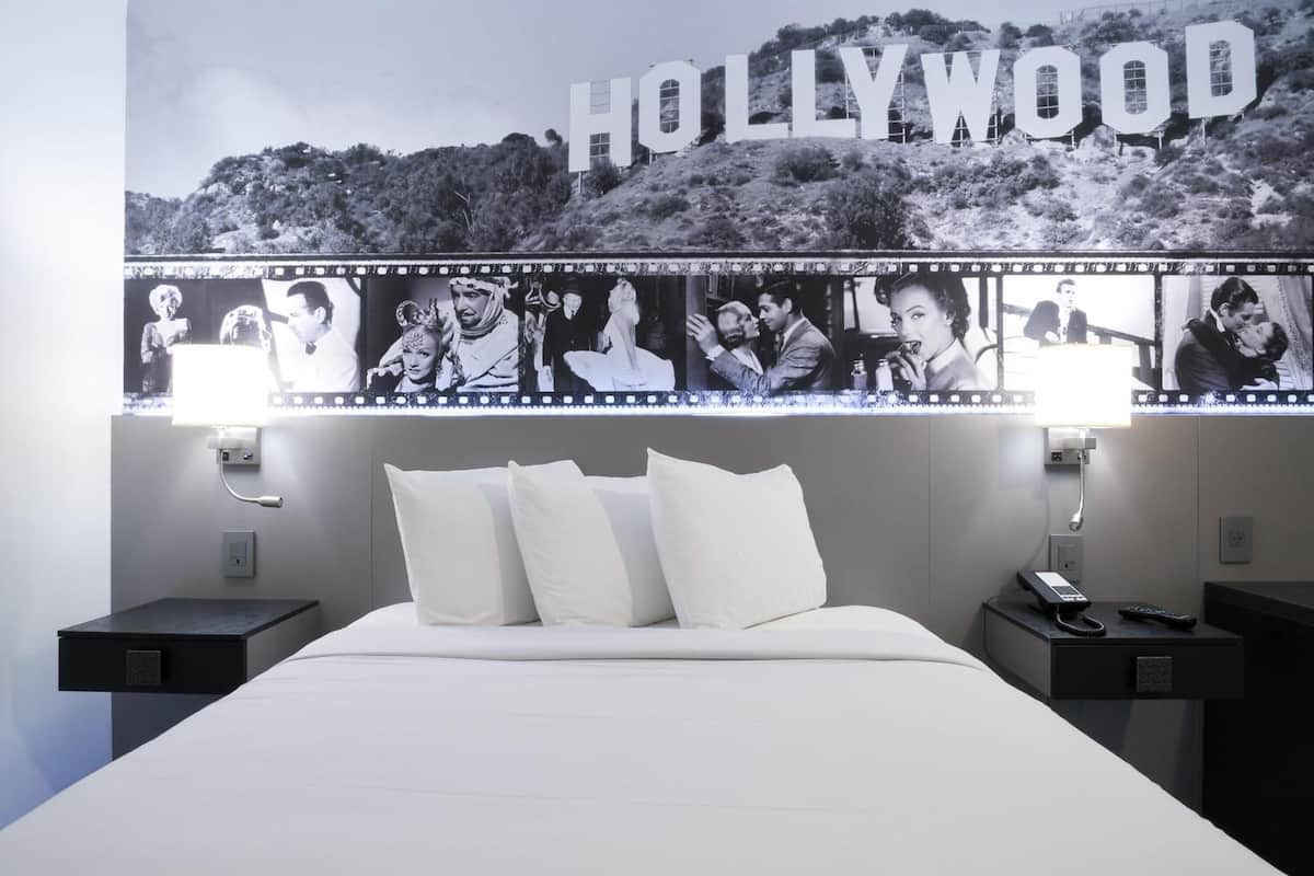 A hotel room with a large Hollywood sign on the wall, a bed with white linens, and two bedside tables with lamps and phones.