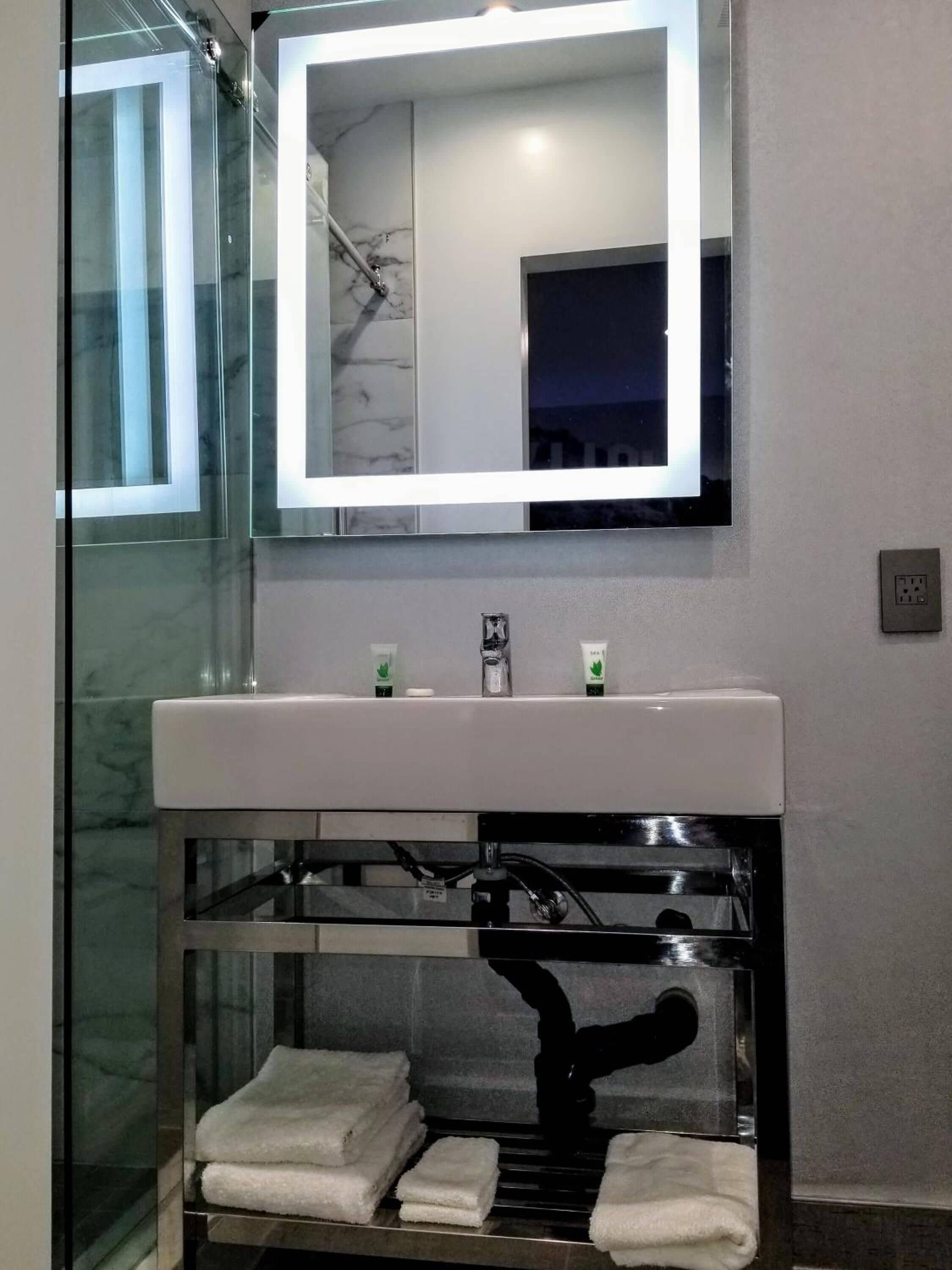 A bathroom with a glass-enclosed shower, a white sink with a marble backsplash, and a mirror with a lighted frame.