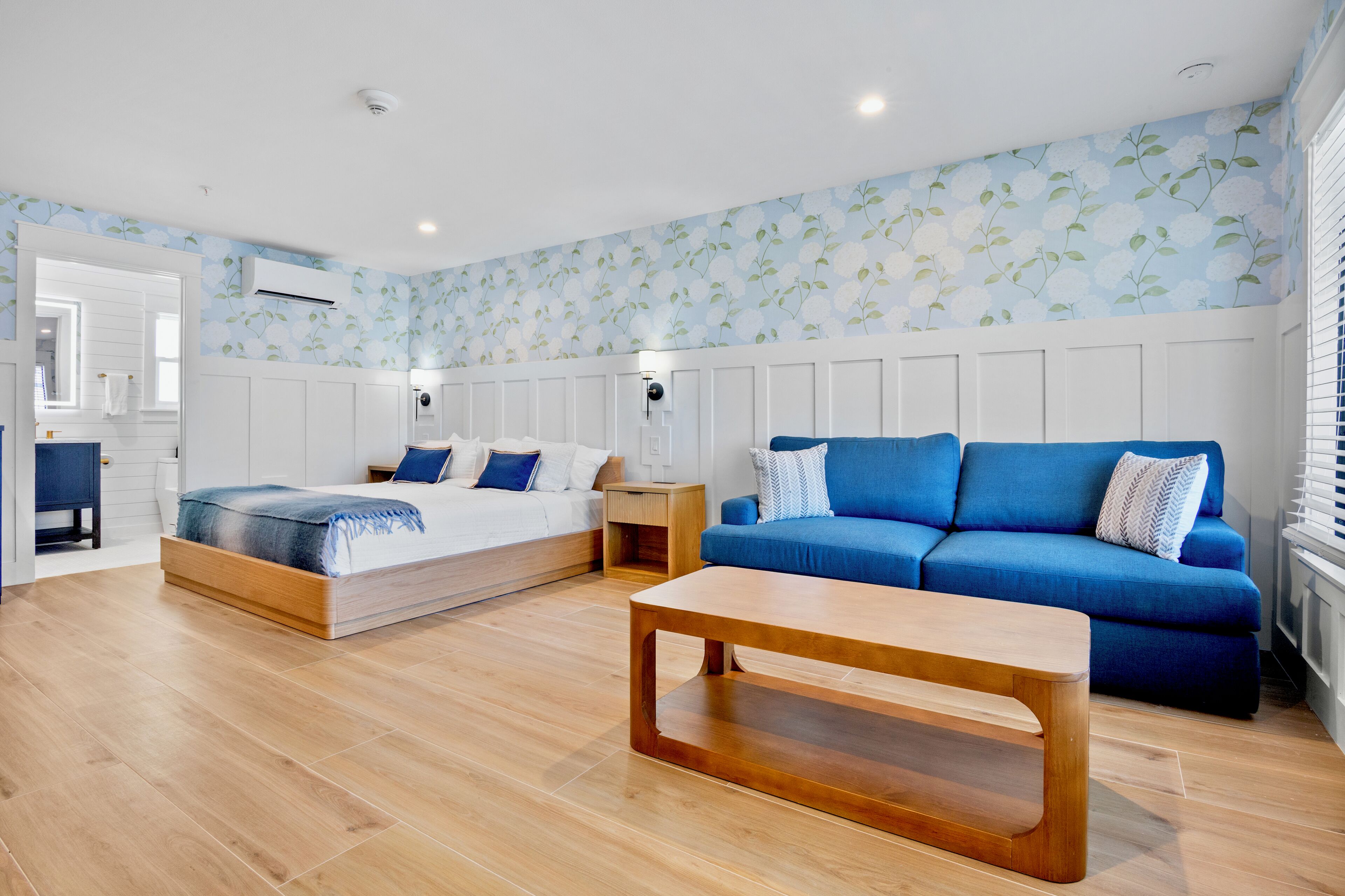 A modern bedroom with a bed, a blue sofa, a wooden coffee table, and a wall with floral wallpaper.