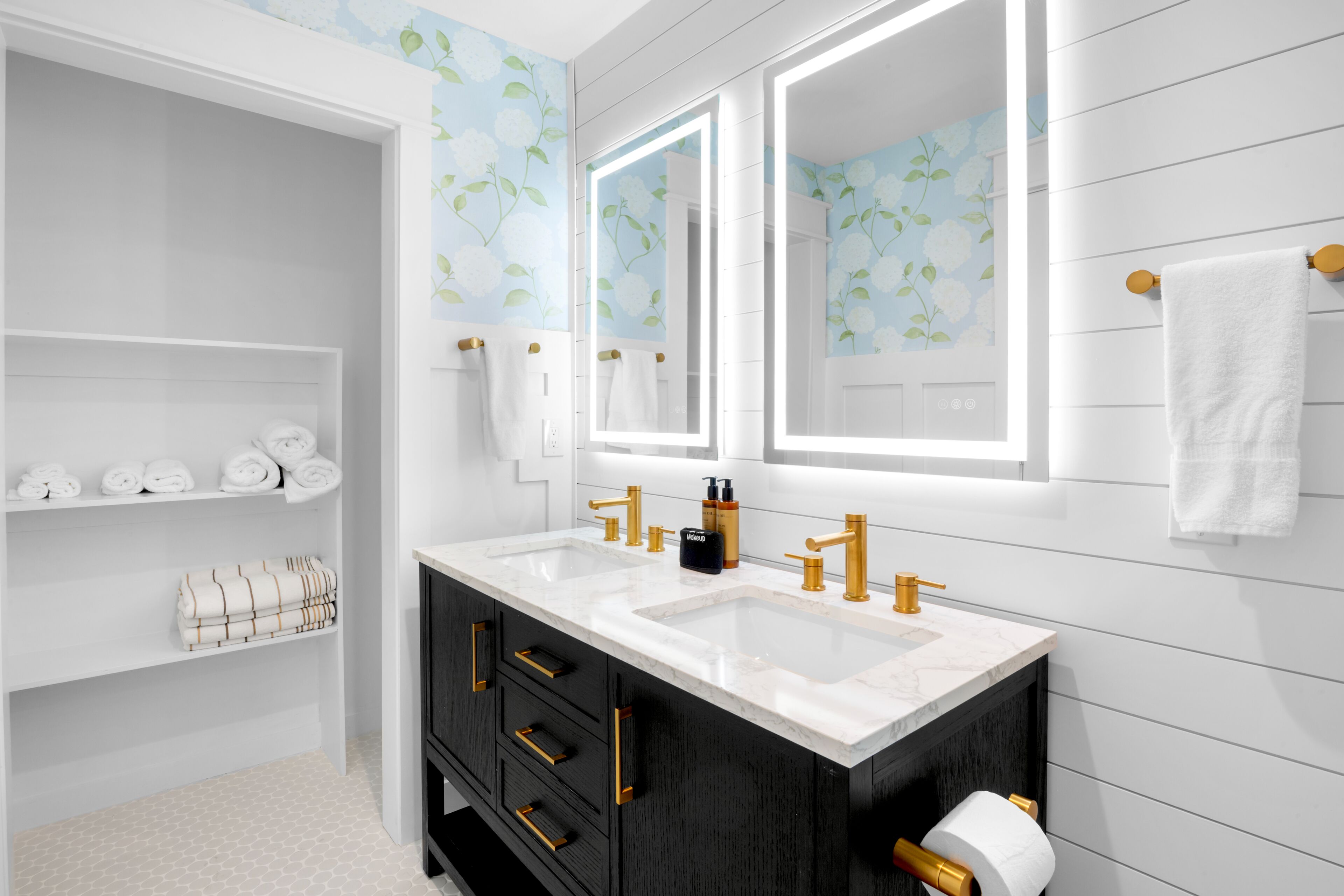 Un bagno con doppio lavabo, mobile contenitore scuro, mensole bianche con asciugamani e carta da parati a motivi floreali.