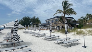 A beach with lounge chairs and striped umbrellas, a building with a balcony, and palm trees.