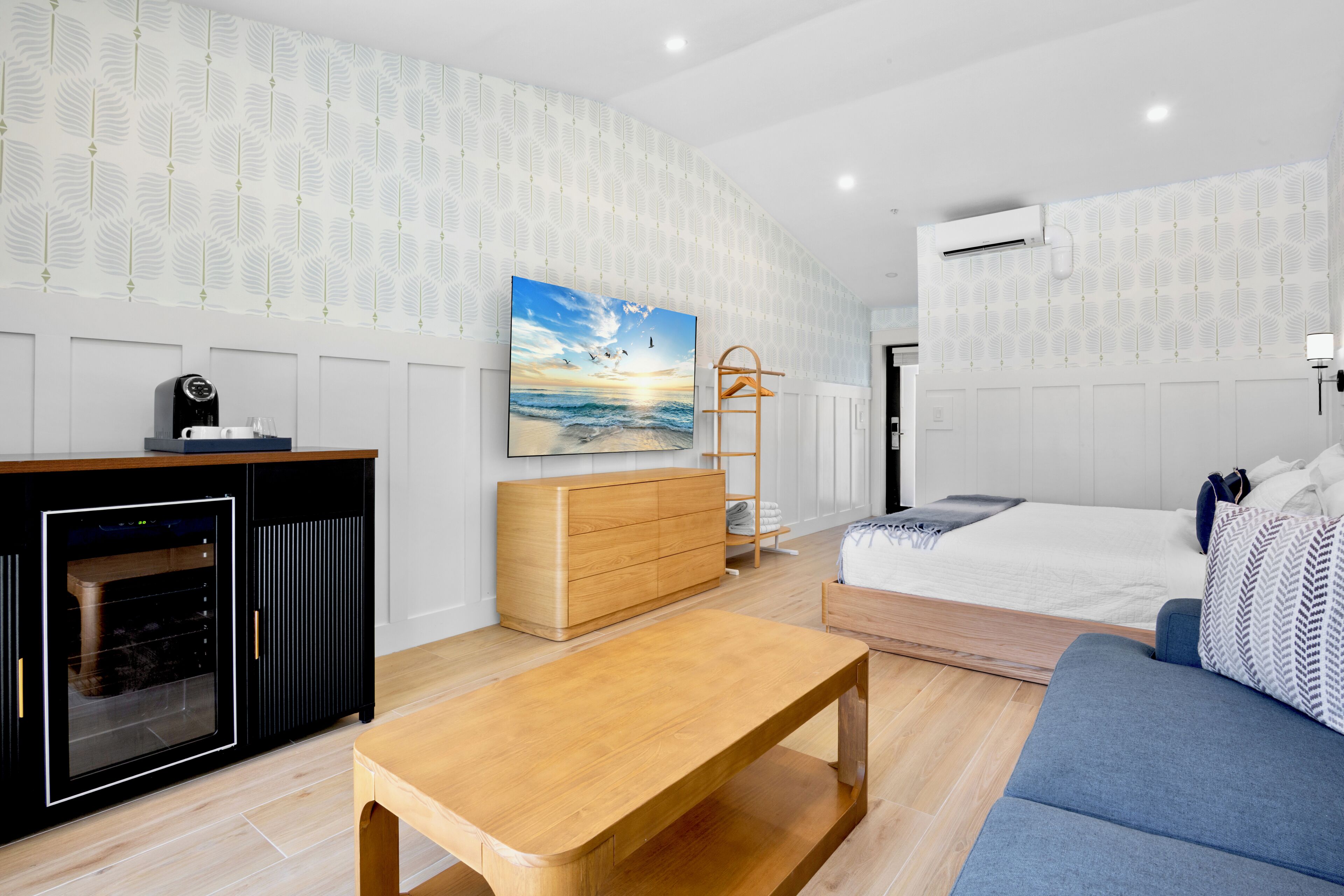 Une chambre d’hôtel moderne avec une table basse en bois, un lit recouvert d’une couverture à motifs bleus et blancs, une commode en bois et un téléviseur fixé au mur.