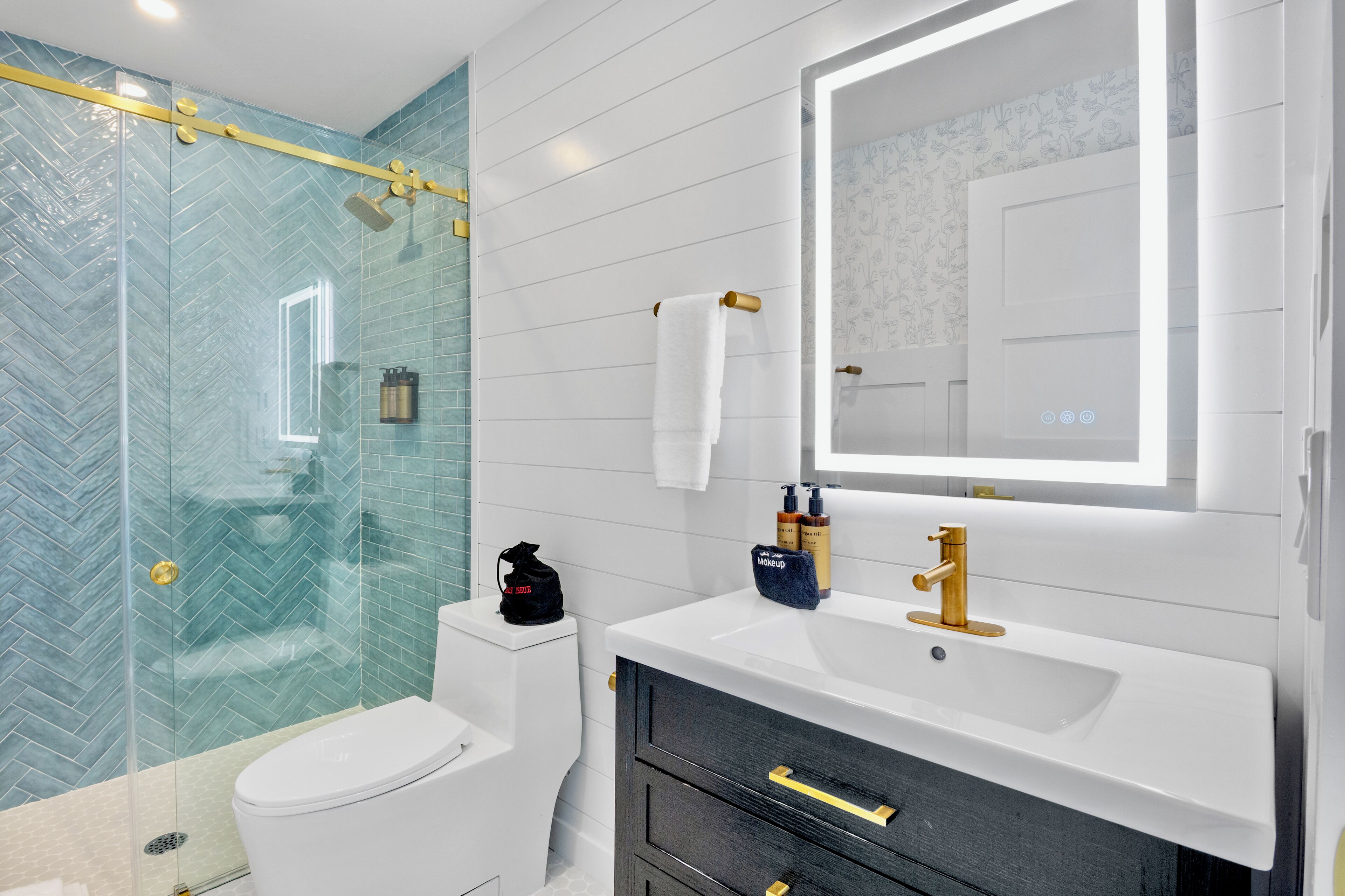 A modern bathroom with a glass shower enclosure, a white toilet, a vanity with a gold faucet, and a mirror with a door.