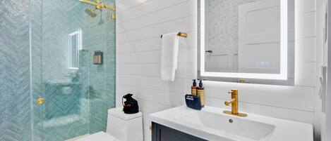 Un baño moderno con cabina de ducha de vidrio, inodoro blanco, tocador con grifo dorado y espejo con puerta.