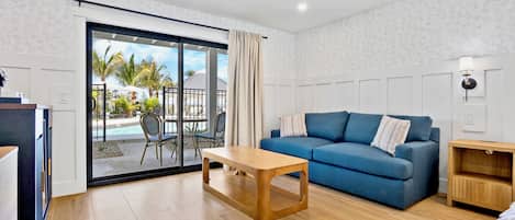 A room with a blue sofa, a wooden coffee table, a sliding glass door leading to a pool area, and a small wooden side table.
