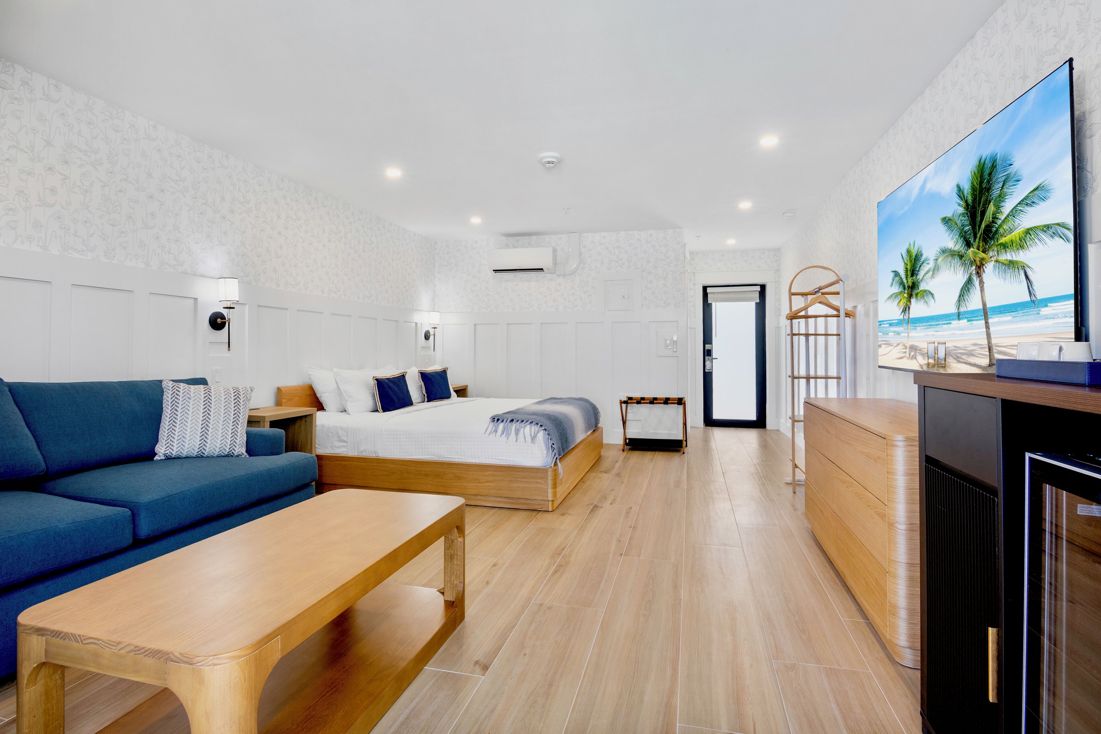 Quarto de hotel moderno com cama, sofá, mesa de centro de madeira e televisão exibindo uma cena de praia.