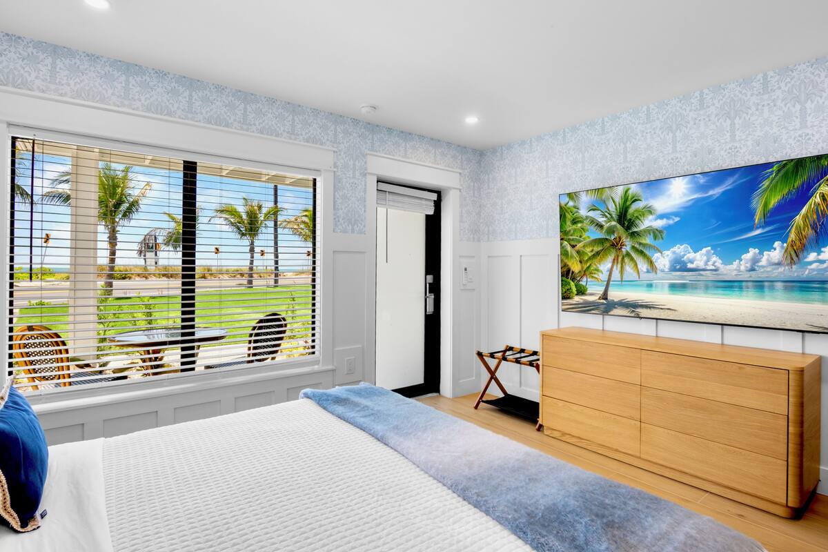 A bedroom with a large bed, a wooden dresser, a TV on the wall, and a window with blinds.