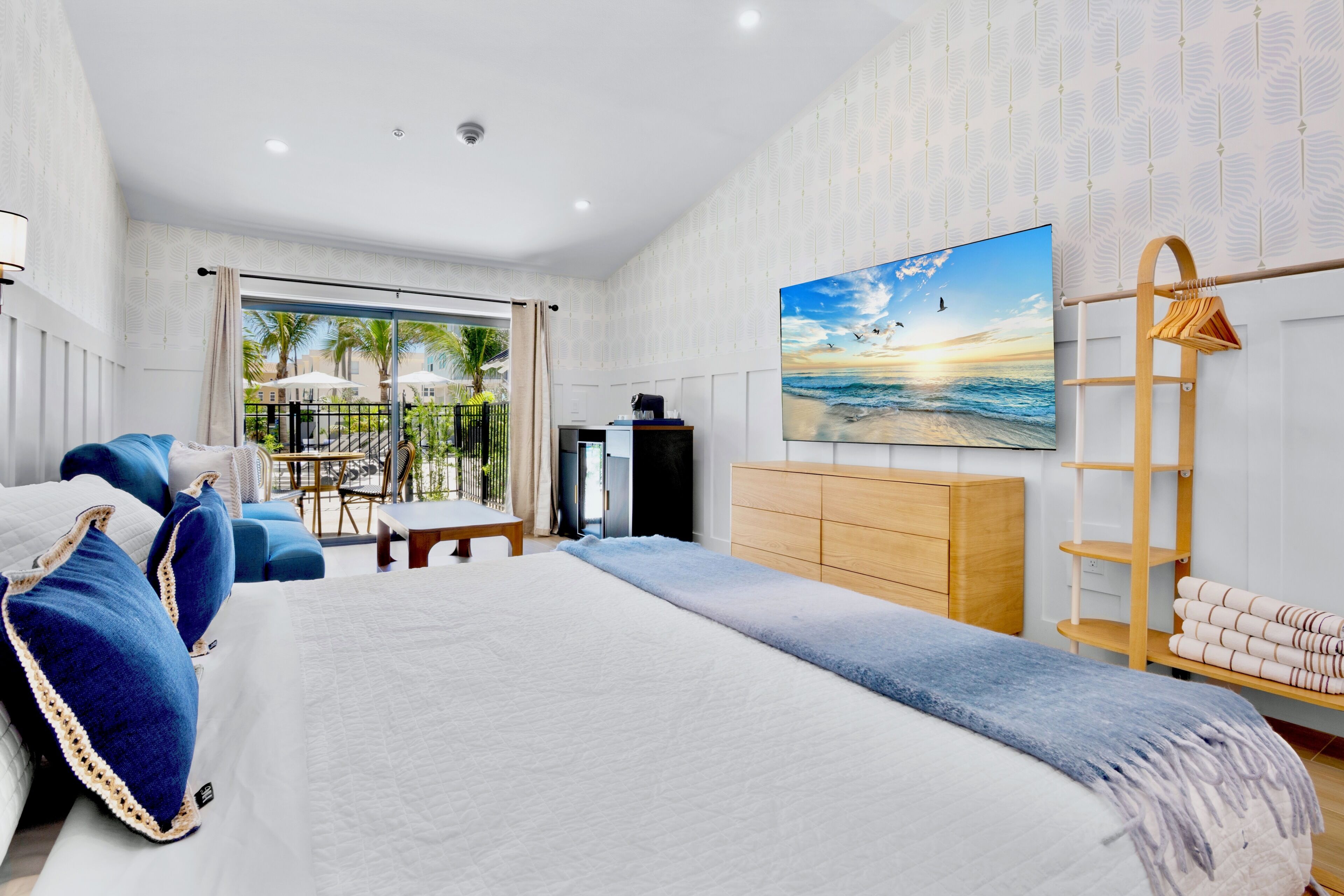 A modern hotel room with a large bed, a TV, a wooden dresser, and a balcony with outdoor furniture.