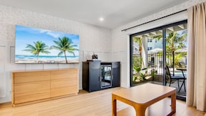 A room with a wooden desk, a TV with a beach scene, a minibar, and a sliding glass door to a balcony with outdoor furniture.