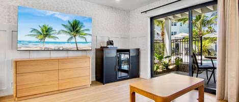 Une chambre dotée d’un bureau en bois, d’un téléviseur diffusant des images de plage, d’un mini-bar et d’une porte-fenêtre coulissante donnant sur un balcon aménagé avec du mobilier d’extérieur.