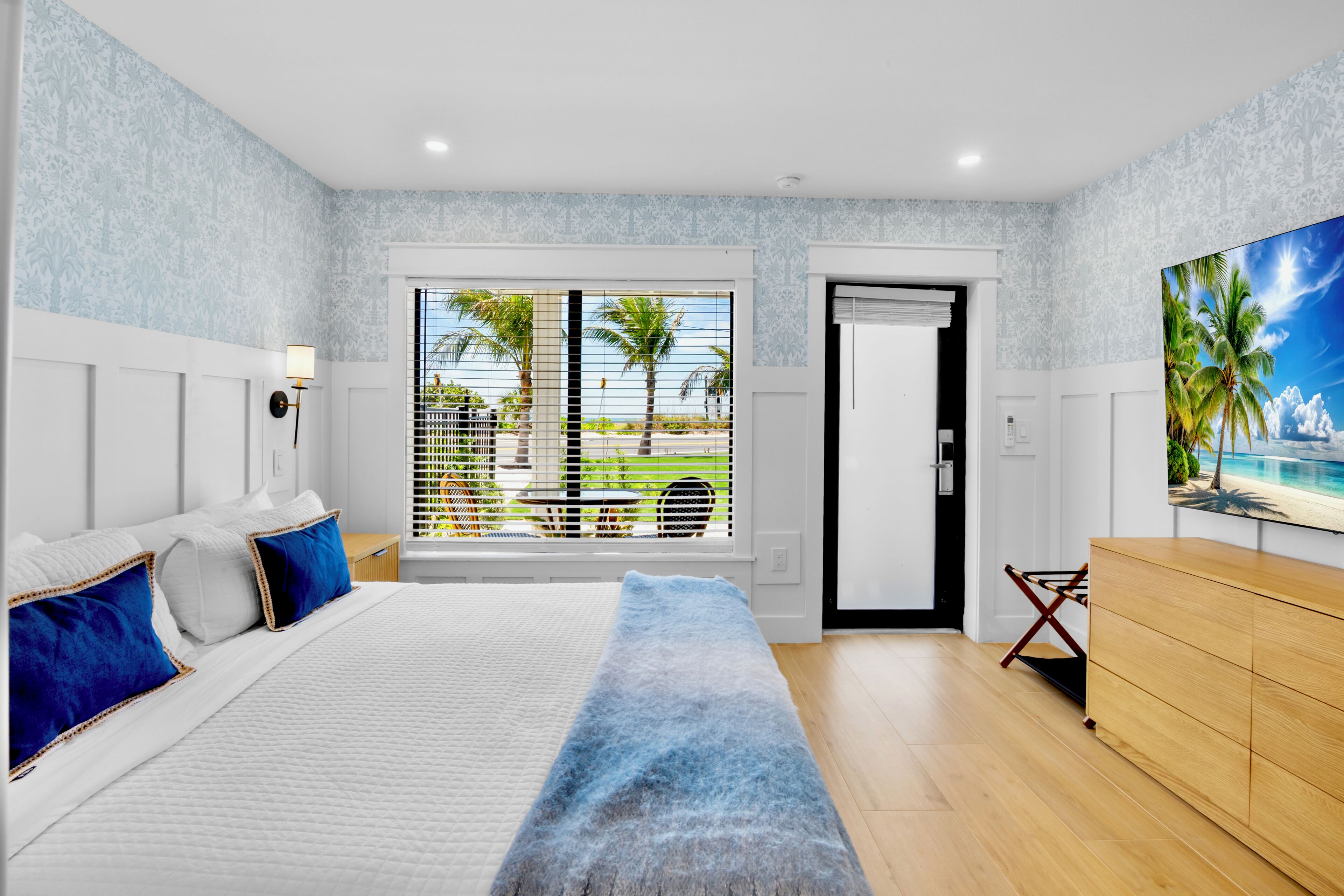 A bedroom with a bed, a dresser, a painting, and a view of palm trees and a pool.