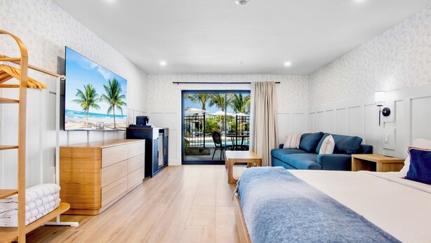 A hotel room with a bed, a sofa, a desk, a TV, a wardrobe, and a balcony with a view of palm trees and the beach.