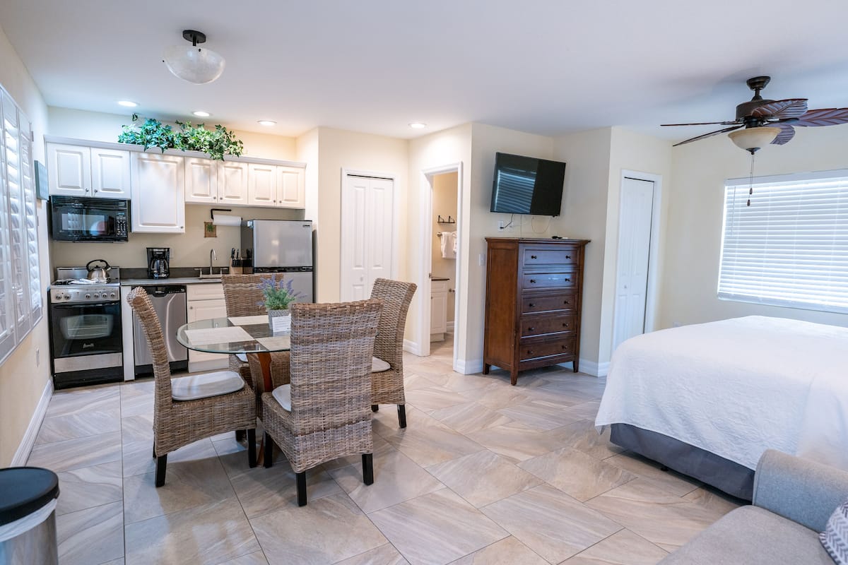 A compact kitchen with modern appliances, a dining area with wicker chairs, a large bed, and a ceiling fan.