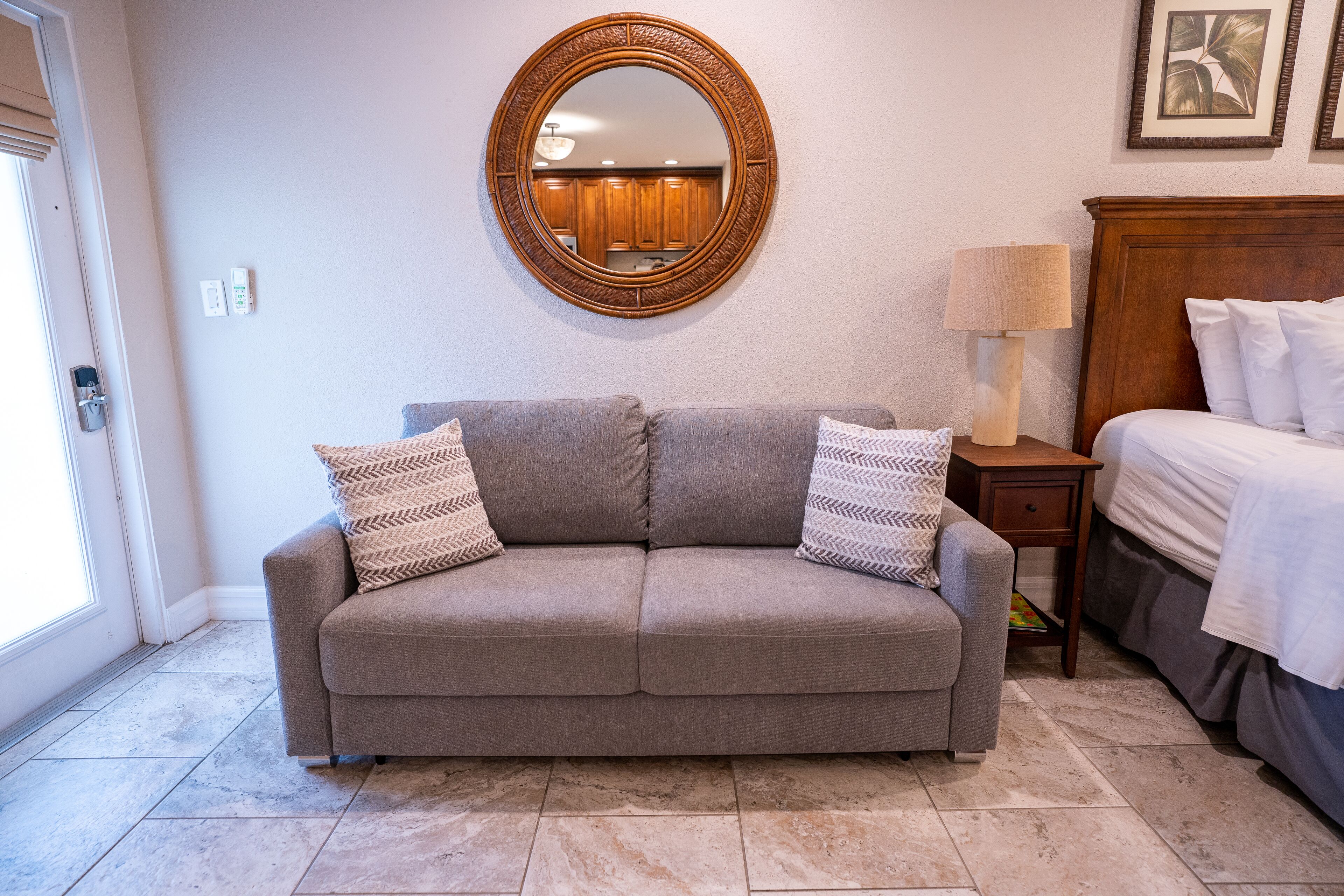 A neutral-toned room with a gray sofa, two patterned pillows, a wooden-framed mirror, a bedside table with a lamp, and a bed with white linens.
