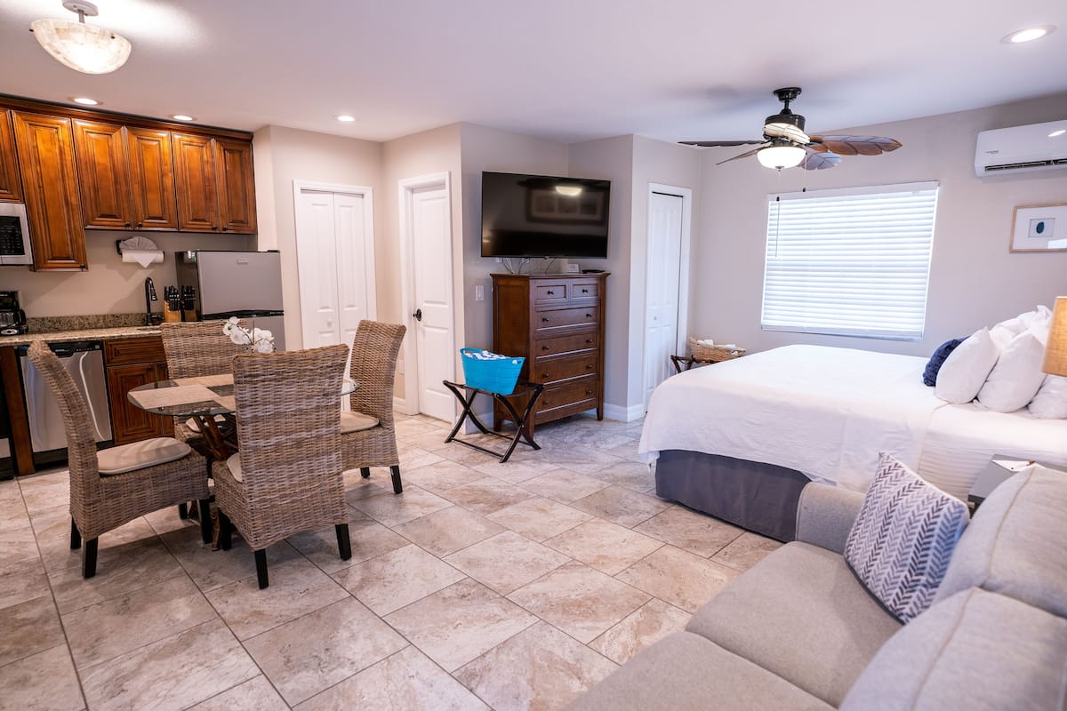 A modern kitchen with wooden cabinets, a dining area with wicker chairs, a bedroom with a large bed and a ceiling fan, and a bathroom with a shower and toilet.