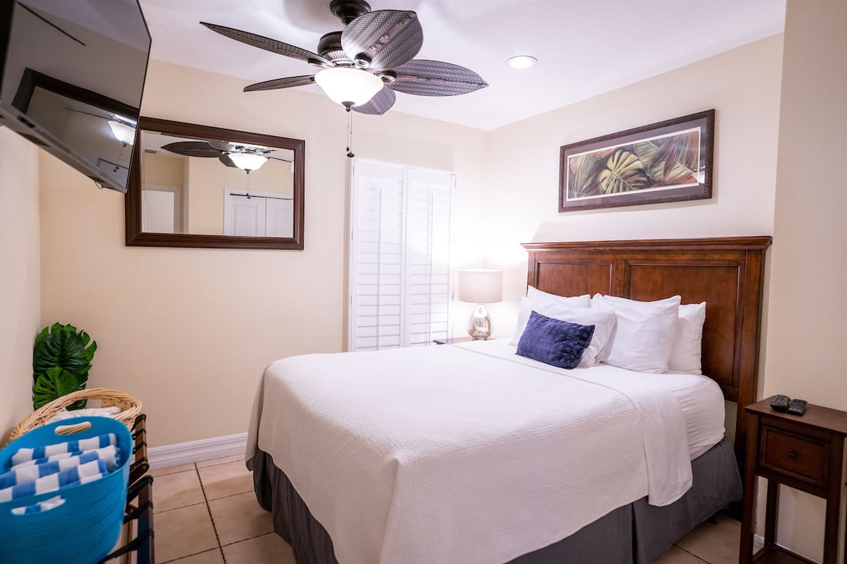 A hotel room with a bed, a ceiling fan, a TV, a mirror, a window with blinds, a lamp, and a framed picture on the wall.