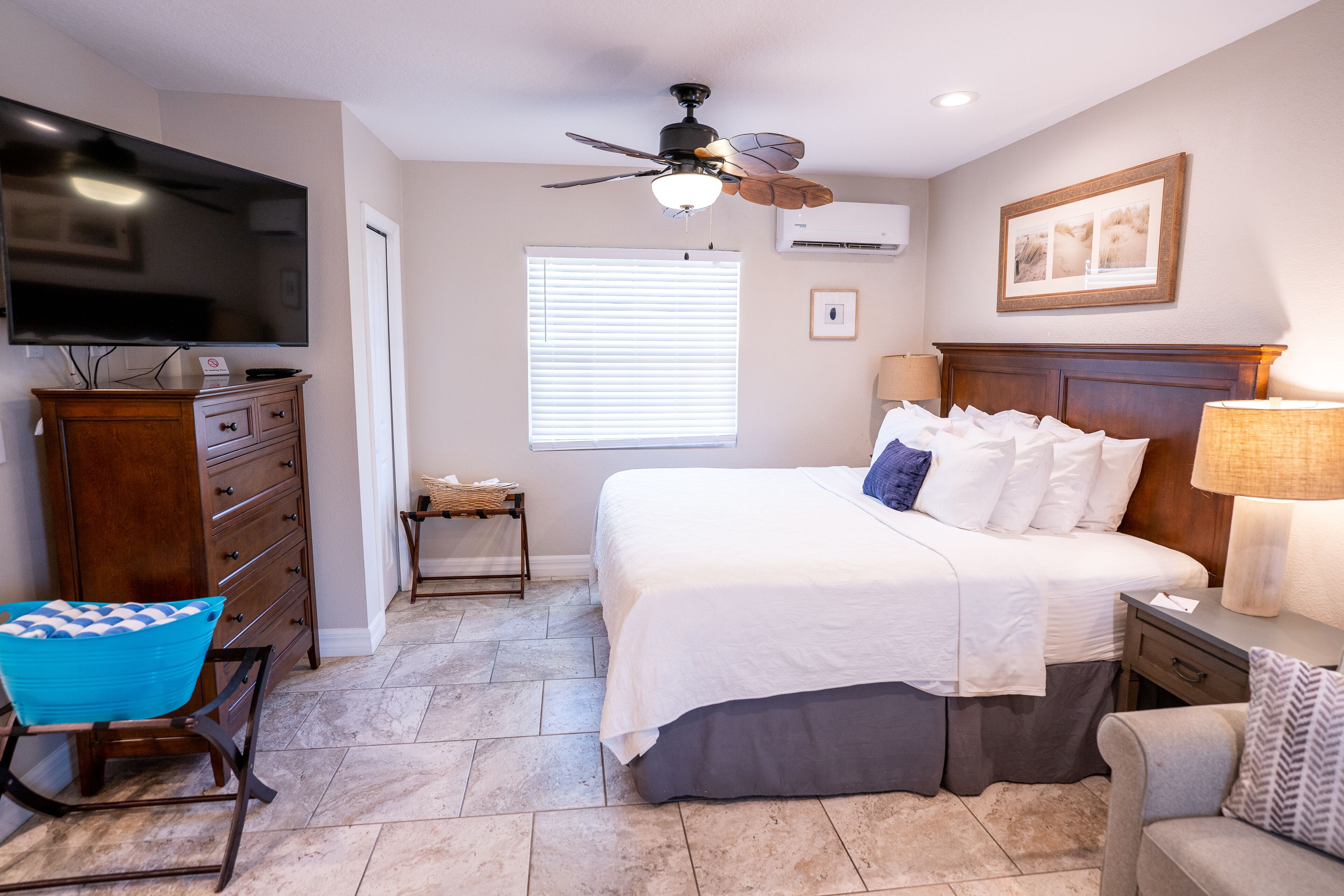A bedroom with a bed, a TV, a ceiling fan, a window with blinds, a nightstand, a lamp, a chair, and a dresser.