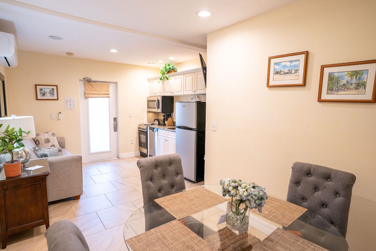 A modern kitchen with white cabinets, a microwave, and a refrigerator. There is a dining table with two chairs and a vase with flowers.