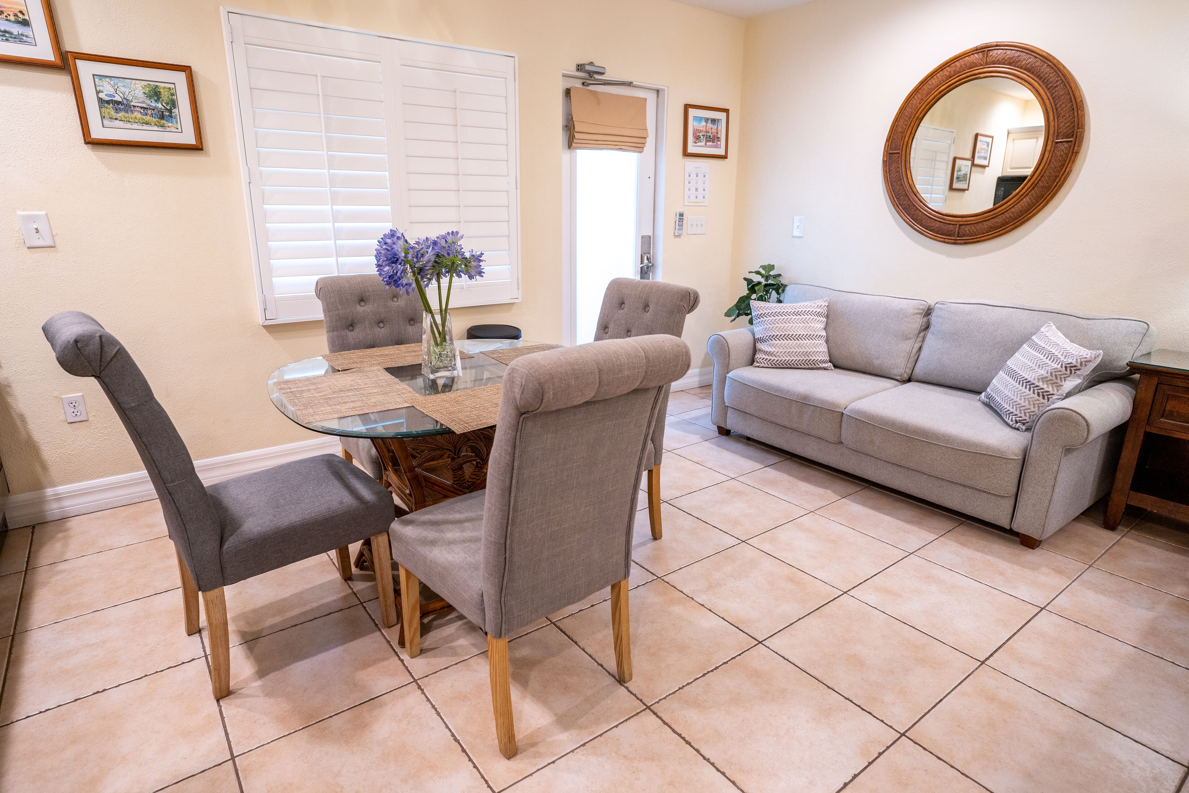 A dining area with a round glass table, four grey upholstered chairs, a vase with purple flowers, and a beige sofa with patterned pillows.