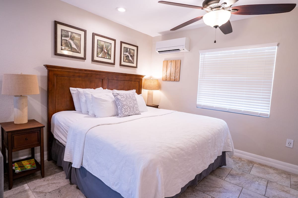 A bedroom with a large bed, two bedside tables with lamps, a ceiling fan, and a window with blinds.