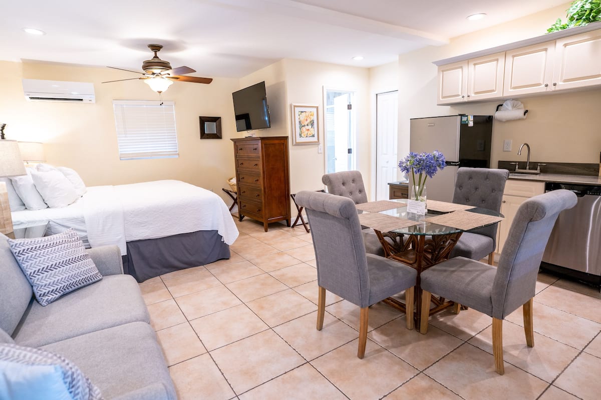 A well-lit room with a bed, a dining table with chairs, a ceiling fan, a TV, and a kitchenette.