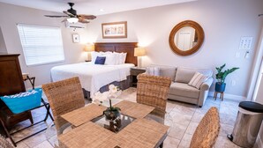 Habitación de hotel con cama, sofá, mesa de comedor, sillas de mimbre, ventilador de techo y planta en maceta.