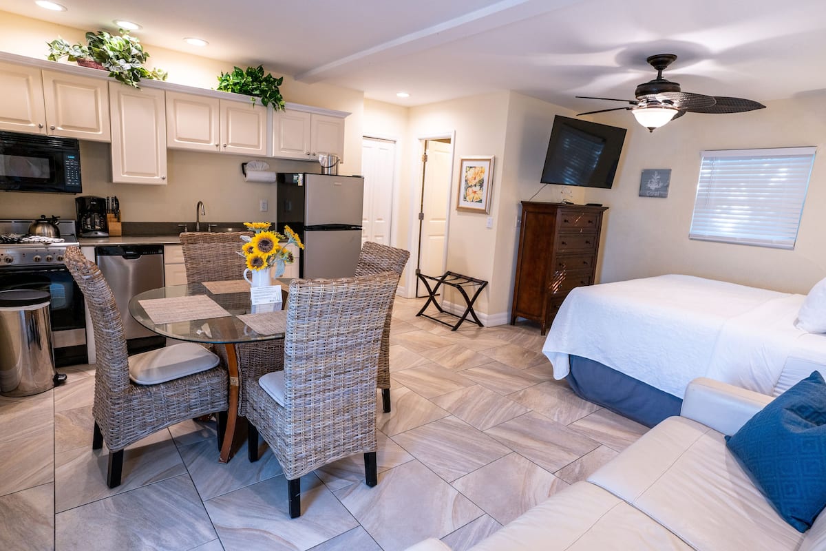 A compact kitchen with white cabinets, a microwave, and a refrigerator. There is a dining area with wicker chairs and a glass table. A bed with white bedding is visible in the background.