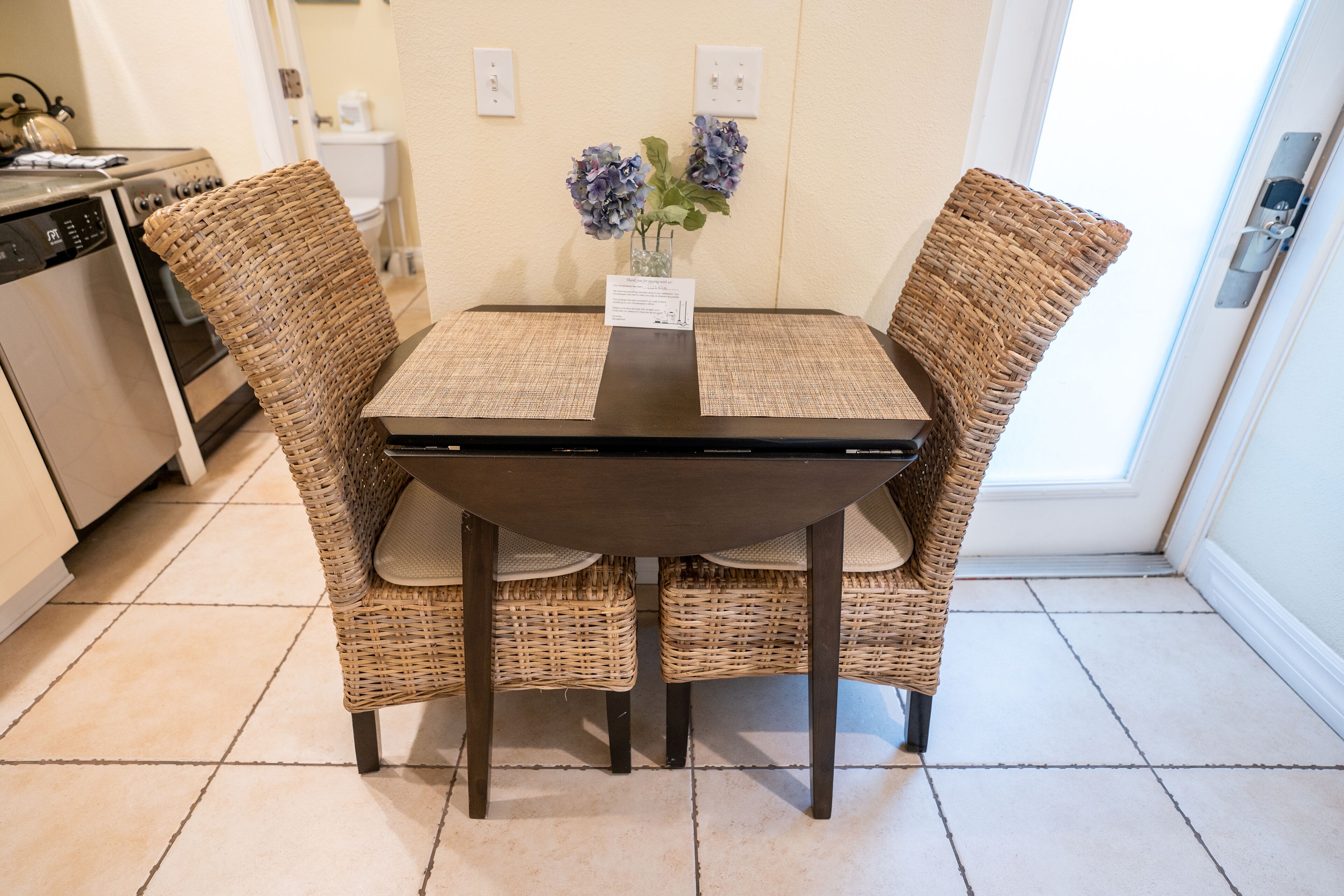 A dining area with a dark wooden table and wicker chairs, a vase with flowers, and a kitchen in the background.