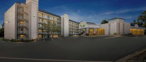 A multi-story hotel with a central entrance, balconies, and a parking lot.