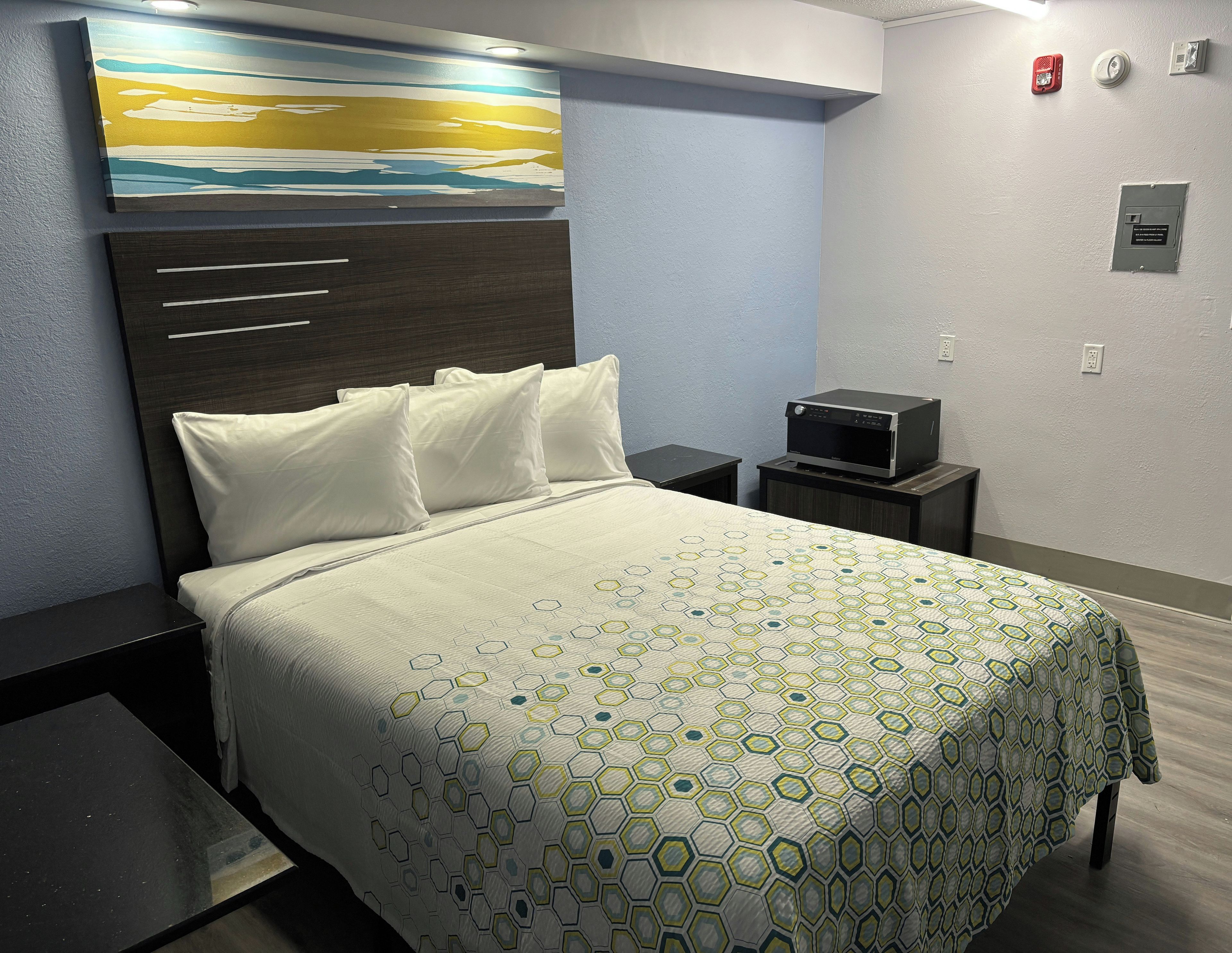 Una habitación de hotel con cama, mesita de noche, televisor montado en la pared y un cuadro sobre la cama.
