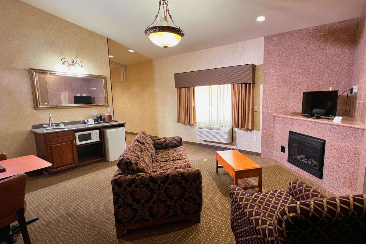 A hotel room with a sofa, armchair, small table, kitchenette, TV, fireplace, and a large mirror.