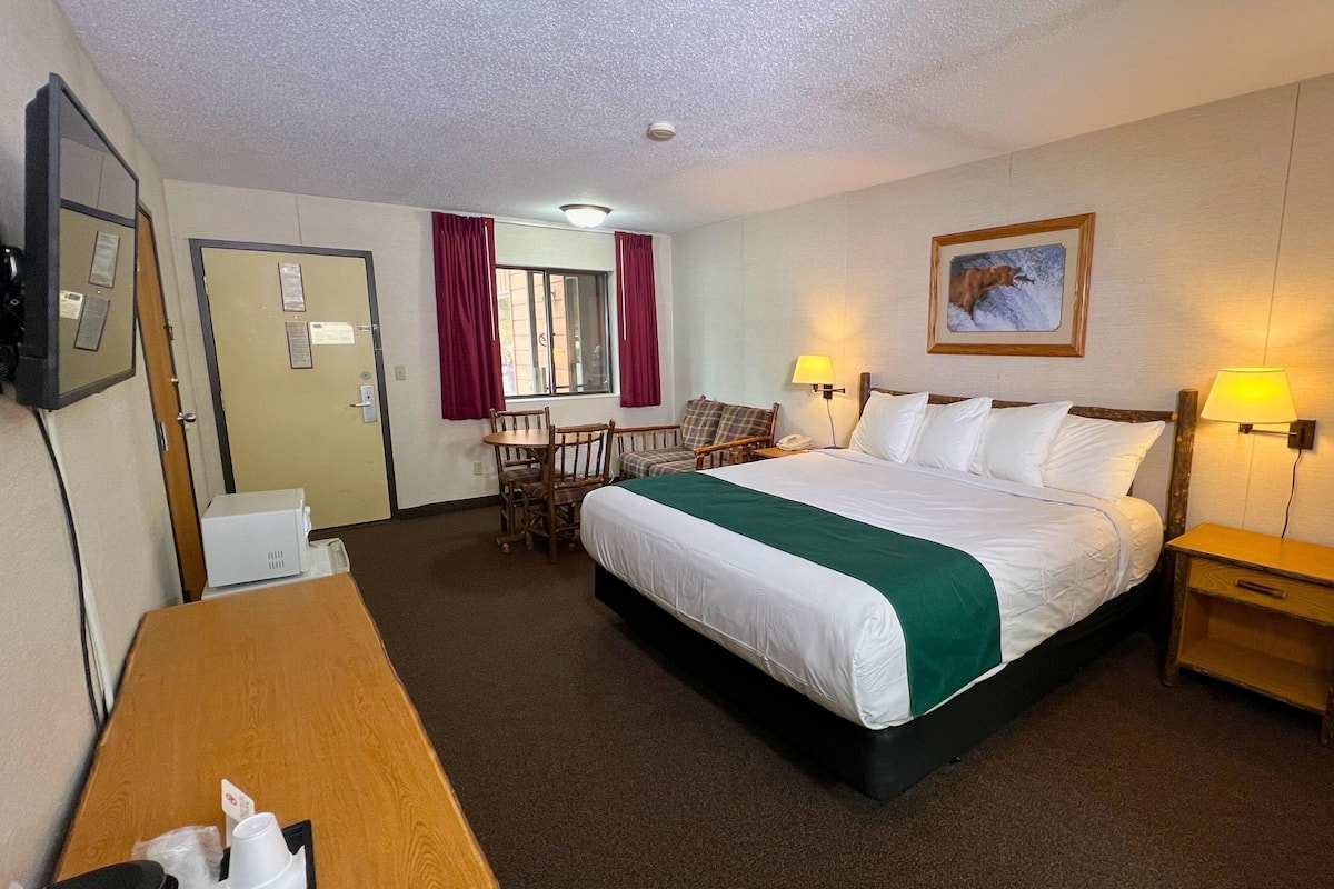 A hotel room with a large bed, two bedside tables with lamps, a desk with a chair, a window with curtains, and a painting on the wall.