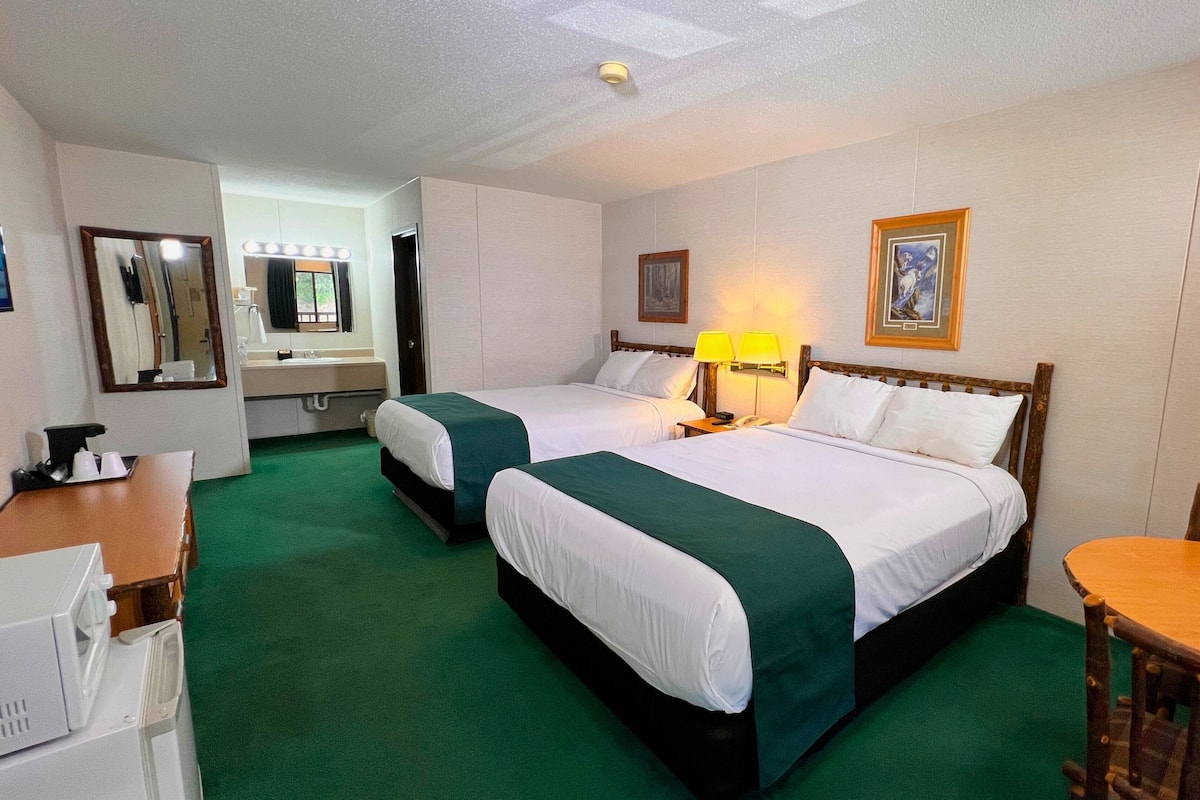 A hotel room with two beds, a desk, a chair, a mirror, a sink, and a painting on the wall.