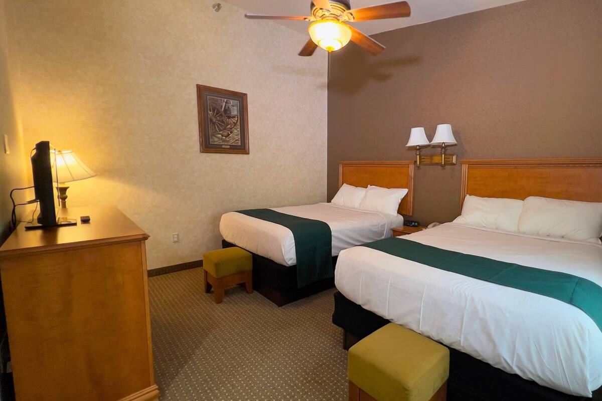 A hotel room with two beds, a desk with a lamp, a ceiling fan, and a wall-mounted picture.