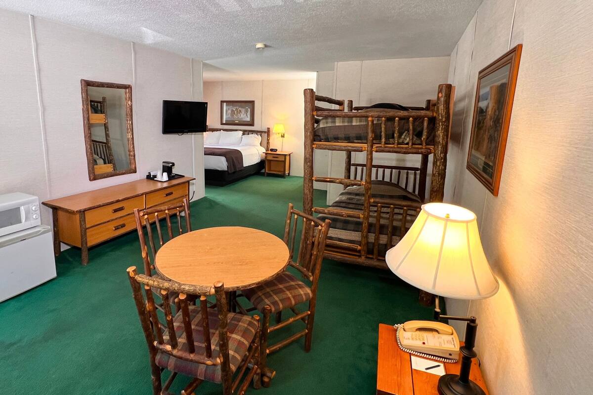 A compact hotel room with a bunk bed, a small desk with a microwave, a round table with chairs, a bedside lamp, and a telephone.