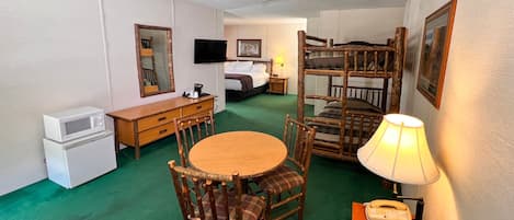 A compact hotel room with a bunk bed, a small desk with a microwave, a round table with chairs, a bedside lamp, and a telephone.