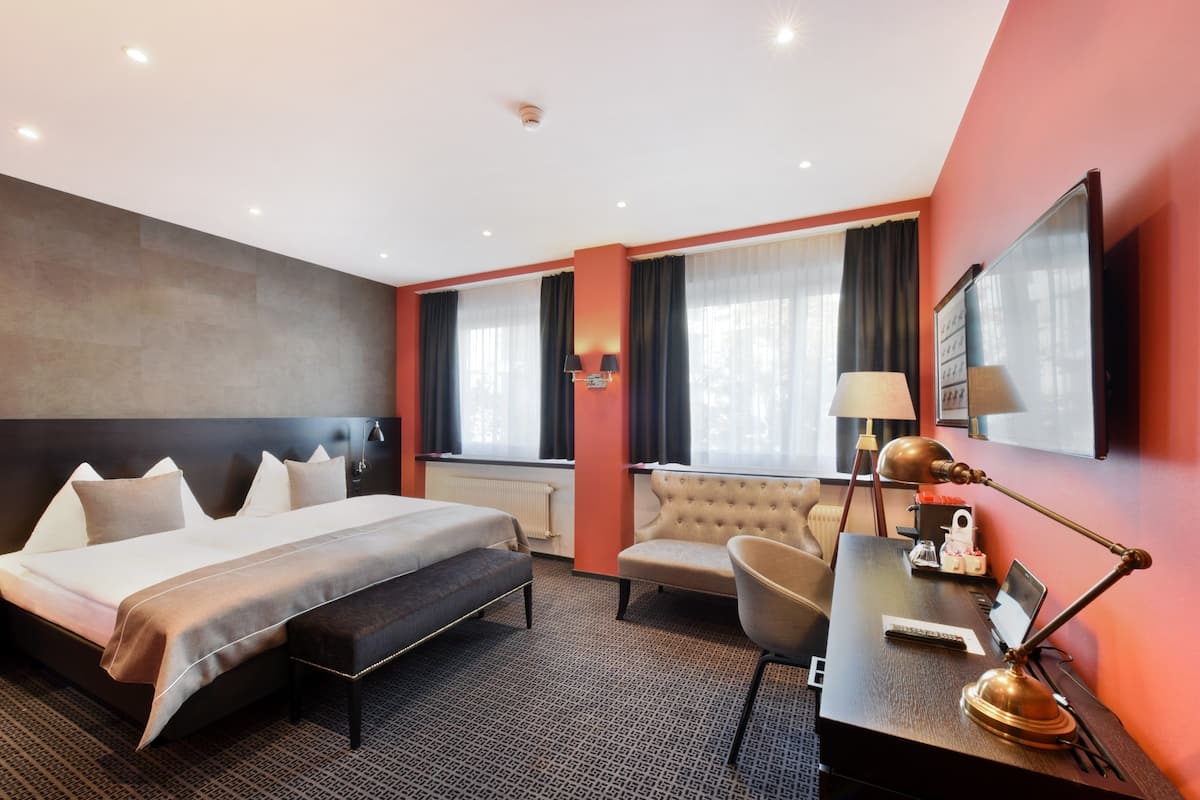 Habitación de hotel con una cama grande, un escritorio con lámpara, una silla y un espejo grande en la pared.