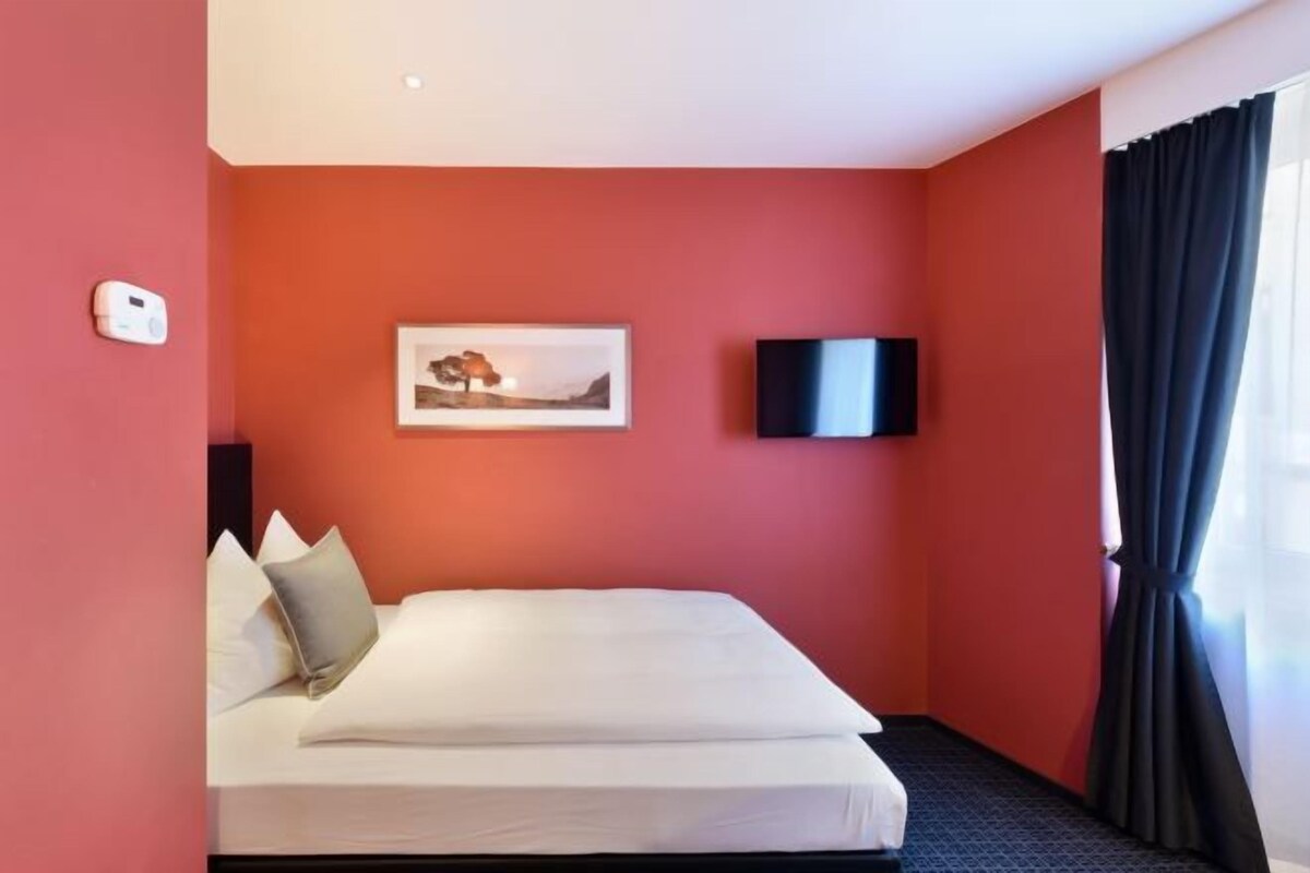 Habitación de hotel con una cama, un cuadro en la pared, un televisor de pantalla plana y una ventana con cortinas.
