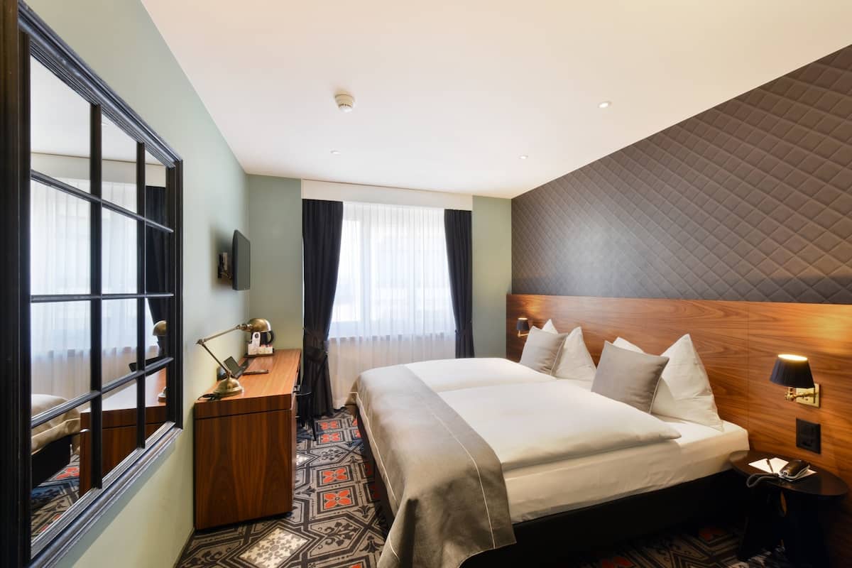 Habitación de hotel con una cama grande, un escritorio con lámpara, armario, televisión y alfombra estampada.