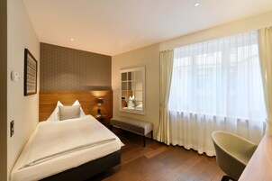 Una habitación de hotel con cama, una banca, una silla, una mesita de noche, un espejo y una ventana con cortinas.