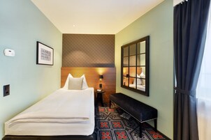 Una habitación de hotel con cama, una banca, un espejo, una lámpara fijada en la pared y una alfombra estampada.