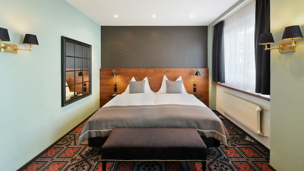 Une chambre d’hôtel avec un grand lit, une tête de lit en bois, une fenêtre avec des rideaux et un sol à motifs.