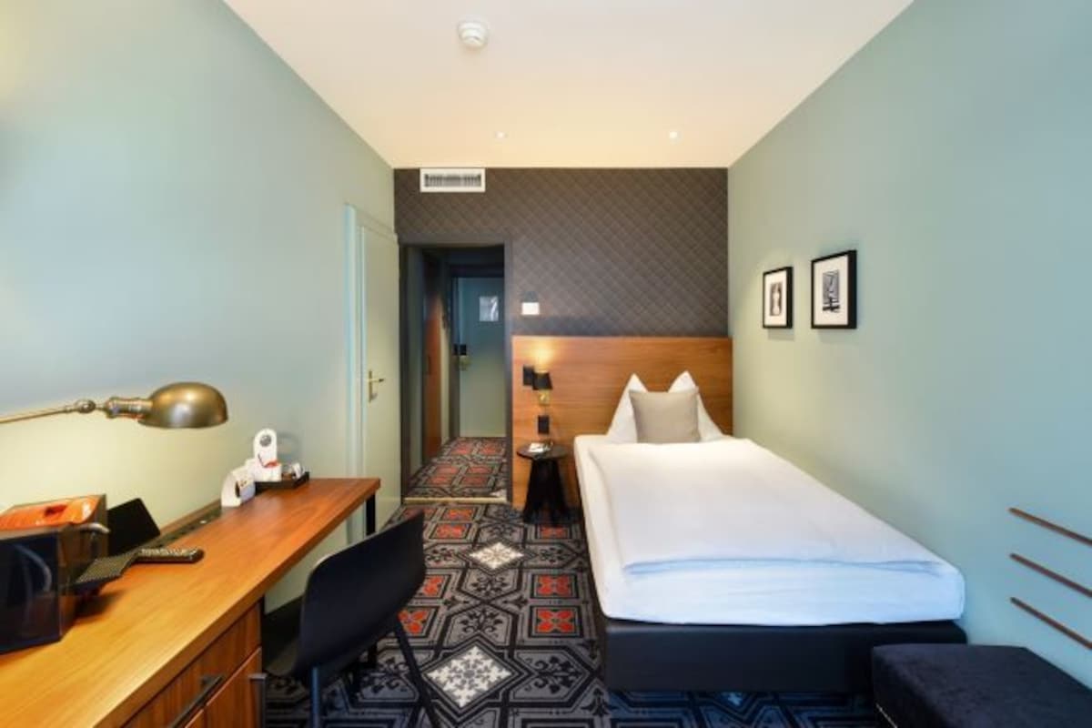 Une chambre d’hôtel avec un bureau en bois, un lit, une petite table avec une lampe et un tapis à motifs.