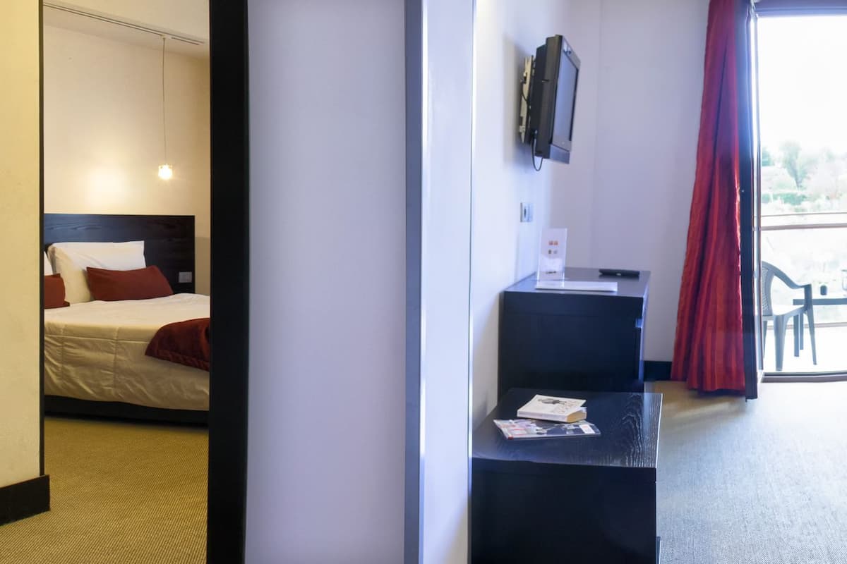 A hotel room with a bed, bedside table, and a small desk with a chair. There is a window with a view of the outside.