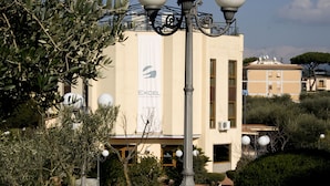 A street lamp with three spherical lights, a building with a beige facade, and a banner with the word "EXCEL". - Excel Hotel Roma Ciampino (Marino)