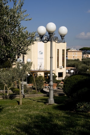 A street lamp with three spherical lights, a building with a beige facade, and a banner with the word "EXCEL". - Excel Hotel Roma Ciampino (Marino)