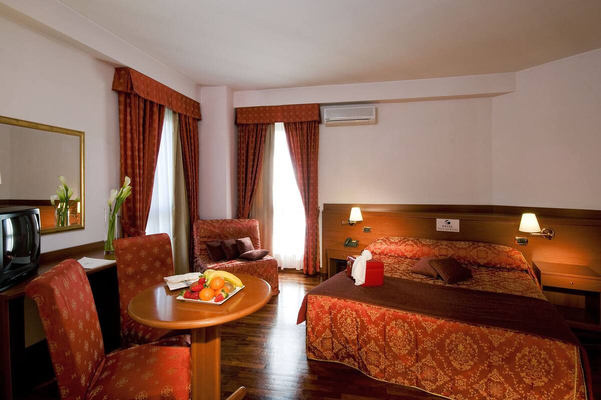 A hotel room with a bed, two chairs, a small table with fruit, a TV, and a mirror.