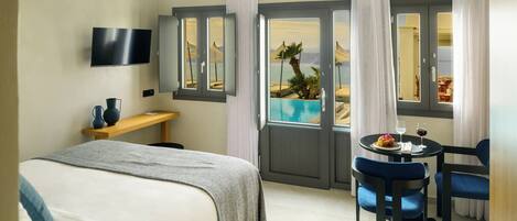 A hotel room with a bed, a desk, a TV, a dining area with a table and chairs, and a view of a pool and palm trees through the door.